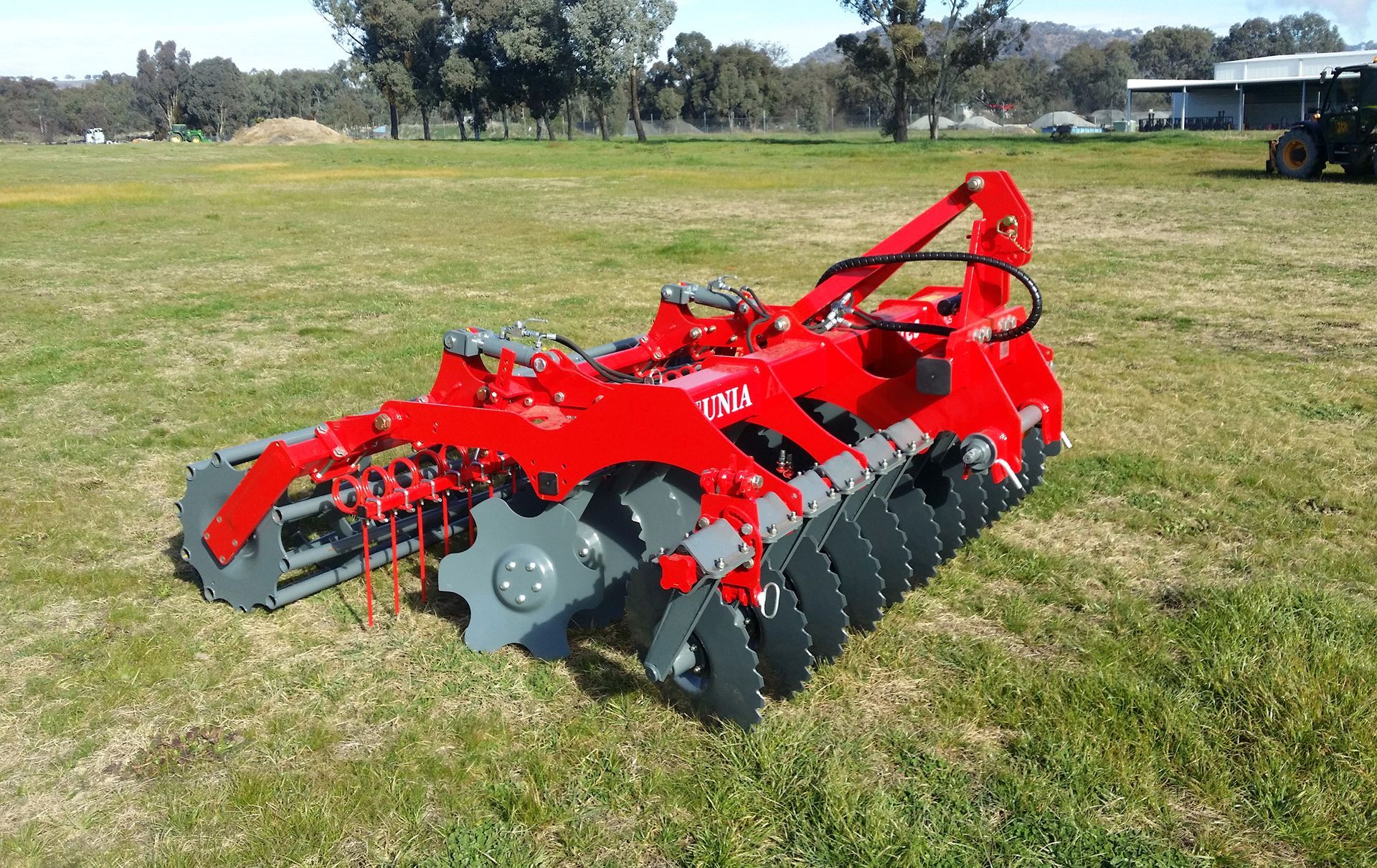A red and gray disc tiller is parked in a grassy field.