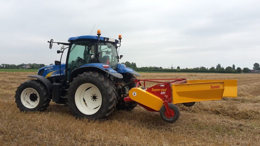 A blue tractor is driving through a field with a yellow trailer attached to it.