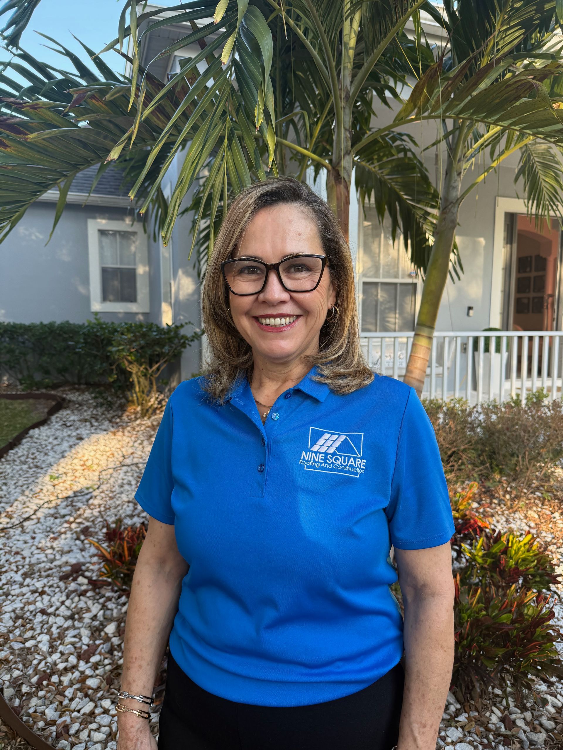 Woman in blue shirt and glasses smiles in front of a house, palm tree behind her.