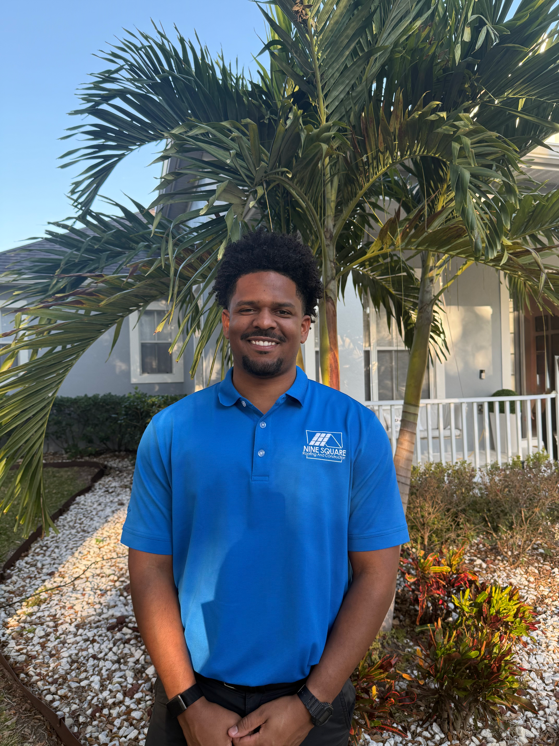 Man in blue polo shirt smiles, stands in front of a house, palm tree, and landscaping.