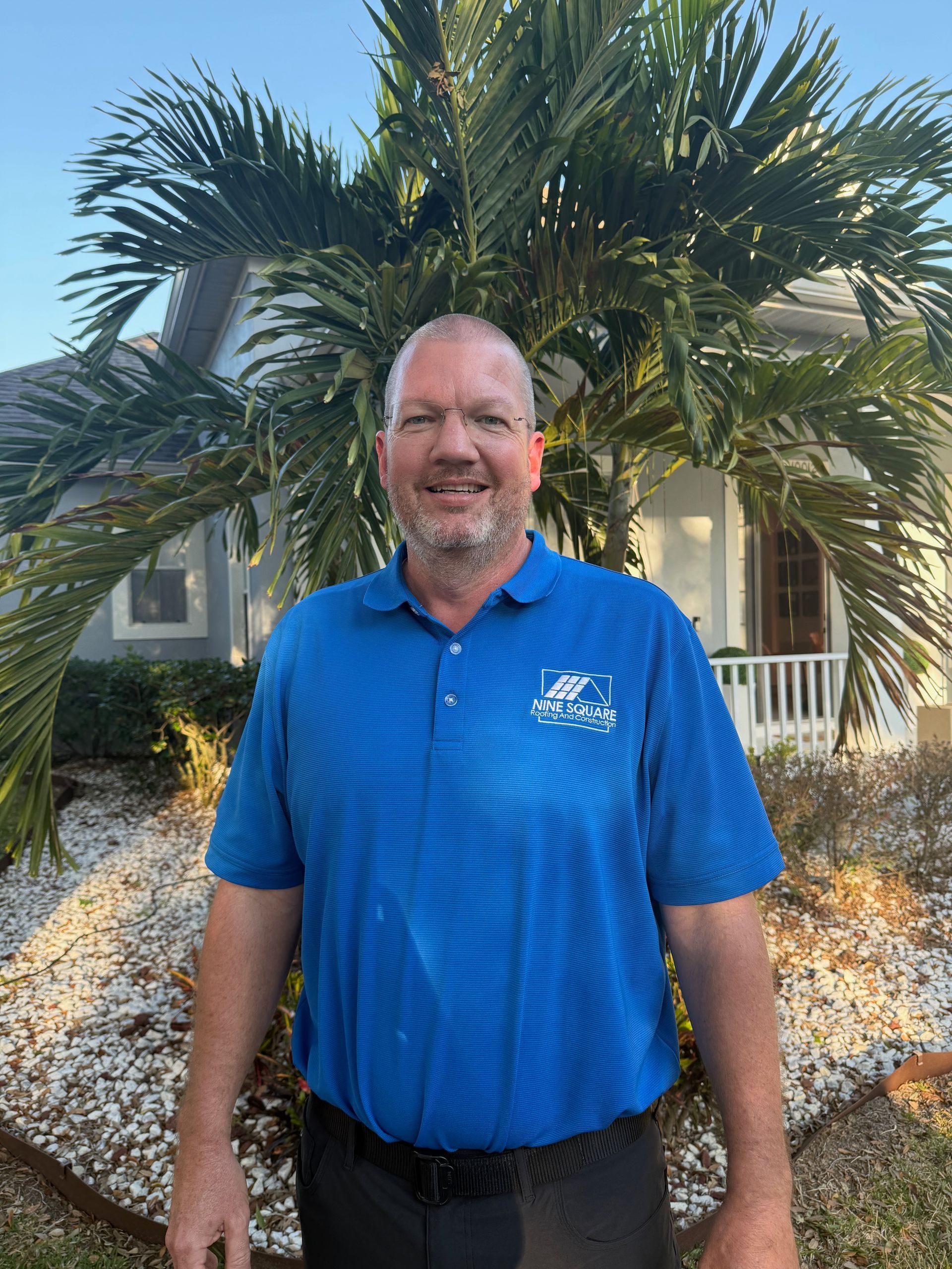 Man wearing blue polo shirt, standing in front of a house and palm tree.