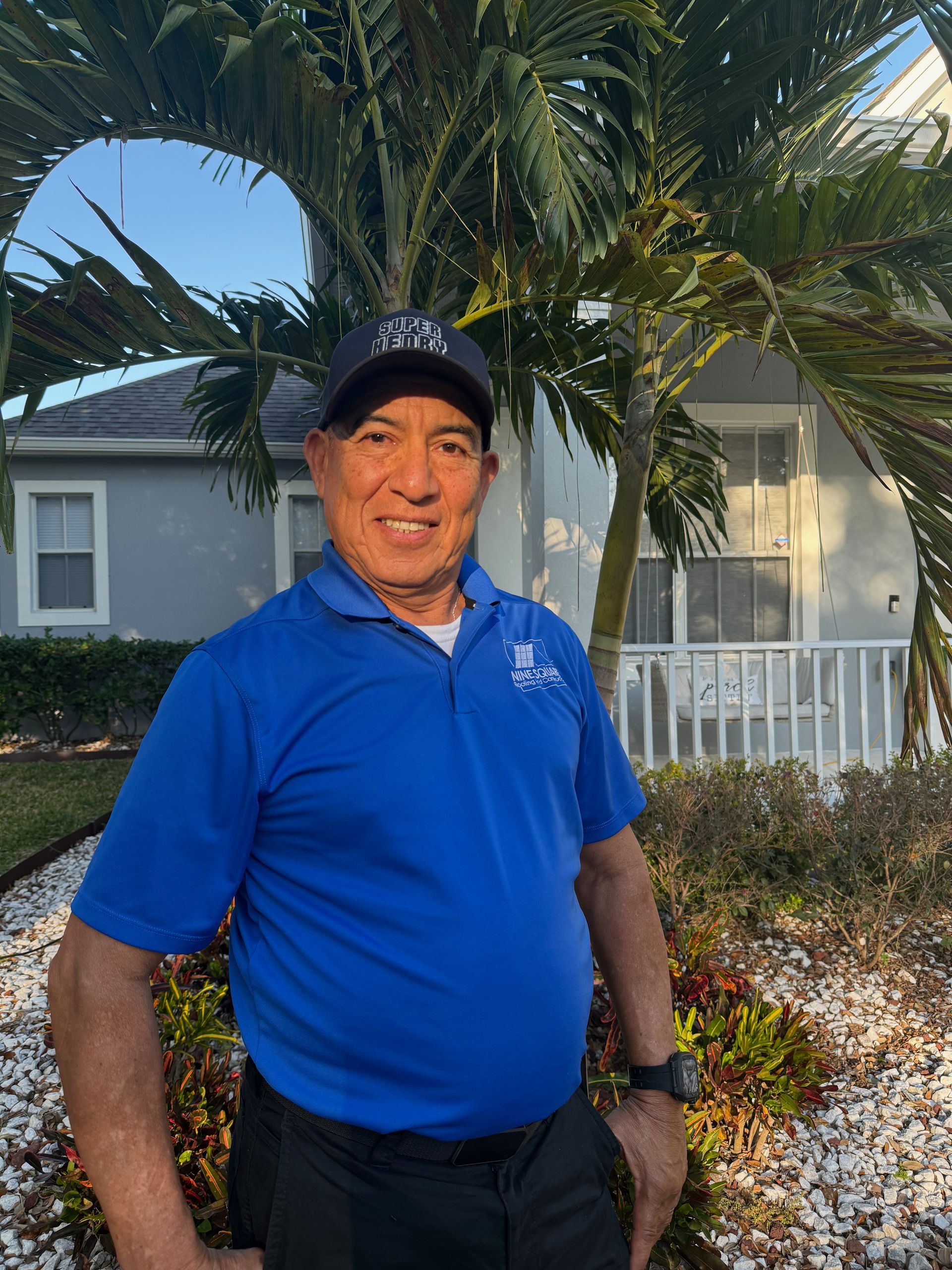 Man in blue polo shirt and cap smiles, standing in front of a house, palm tree in background.