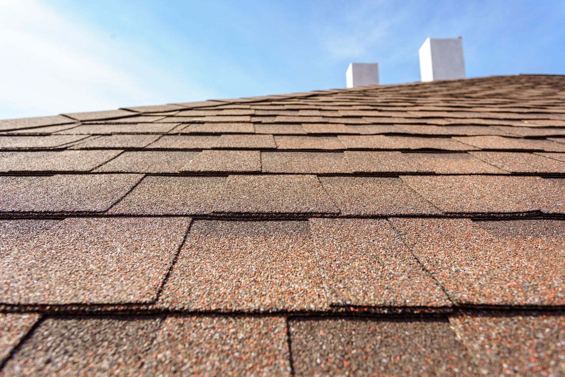 A close up of a roof with shingles and chimneys in the background.