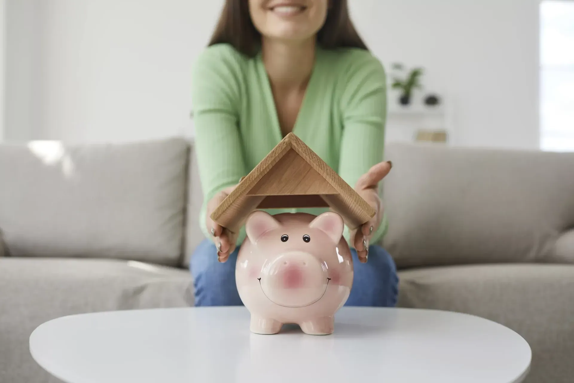 A woman is sitting on a couch holding a piggy bank in the shape of a house.