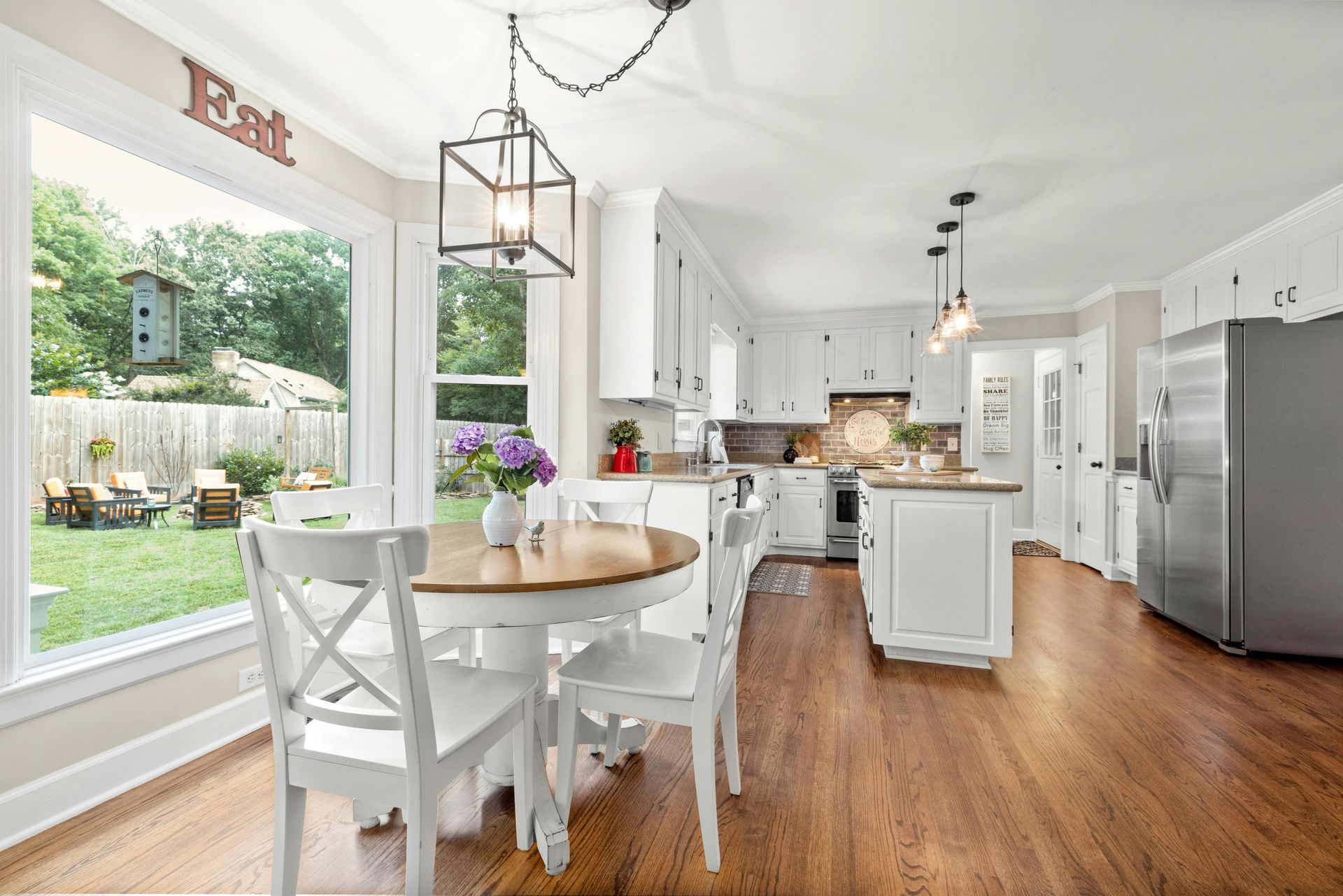 Bright white dining and kitchen area with wood floors, round table, large windows, and stainless steel fridge