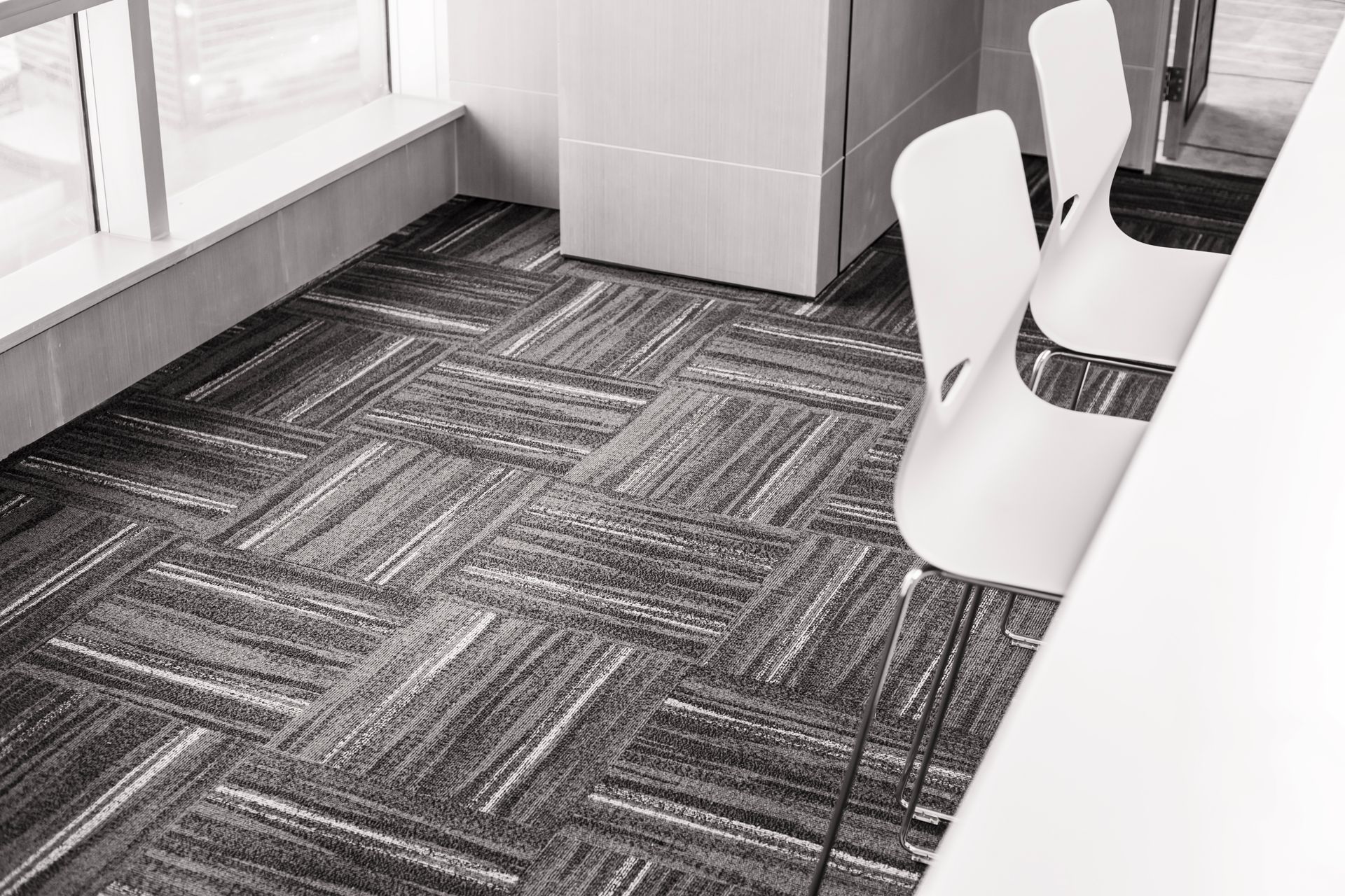 Black and white shot of office space with patterned carpet, white chairs, and a window.