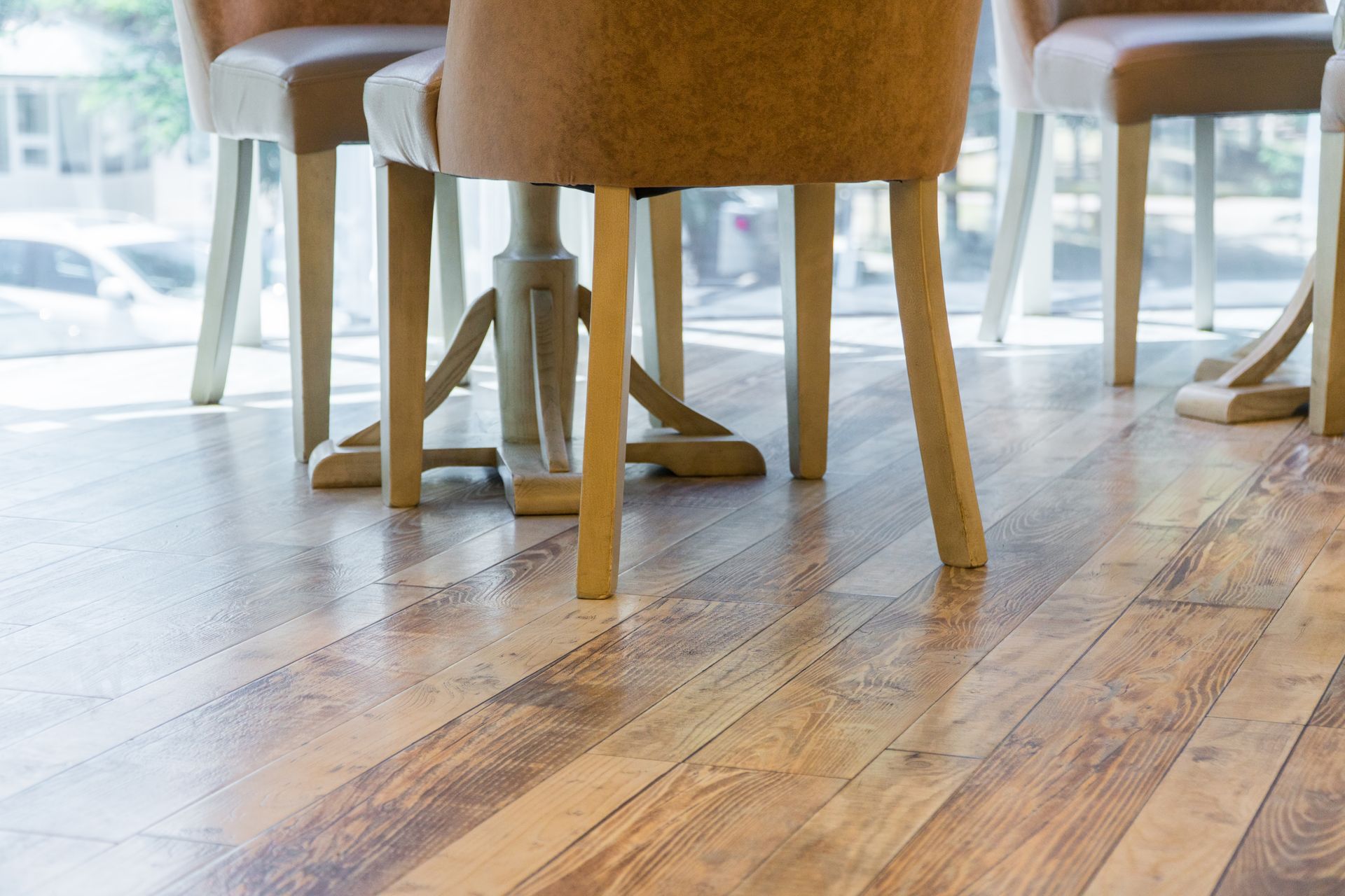 Wooden floor in a bright room with chairs around a table, near a window with a blurred outside view.