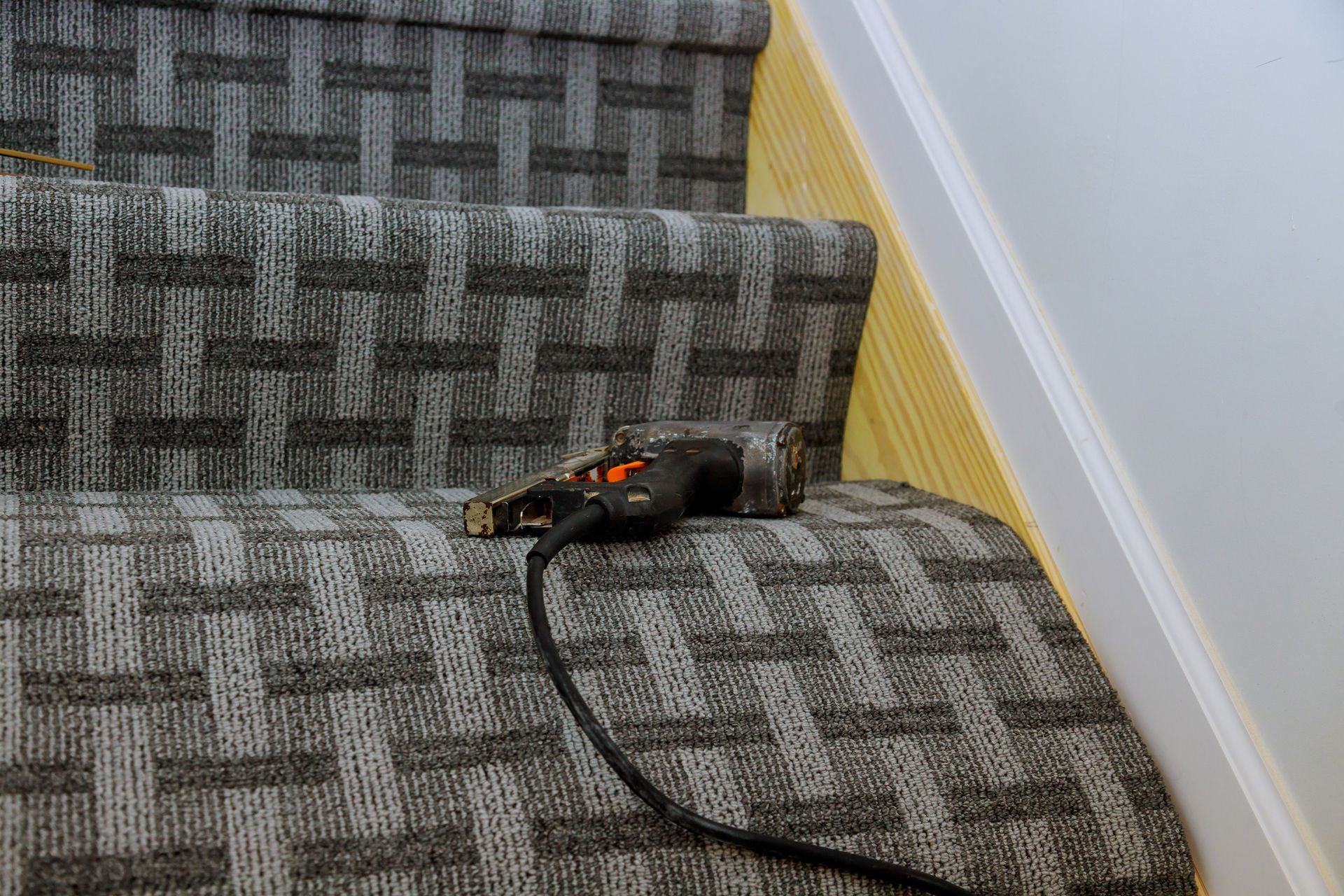 Staircase with patterned gray carpet, a black power tool rests on a step, cord extends.