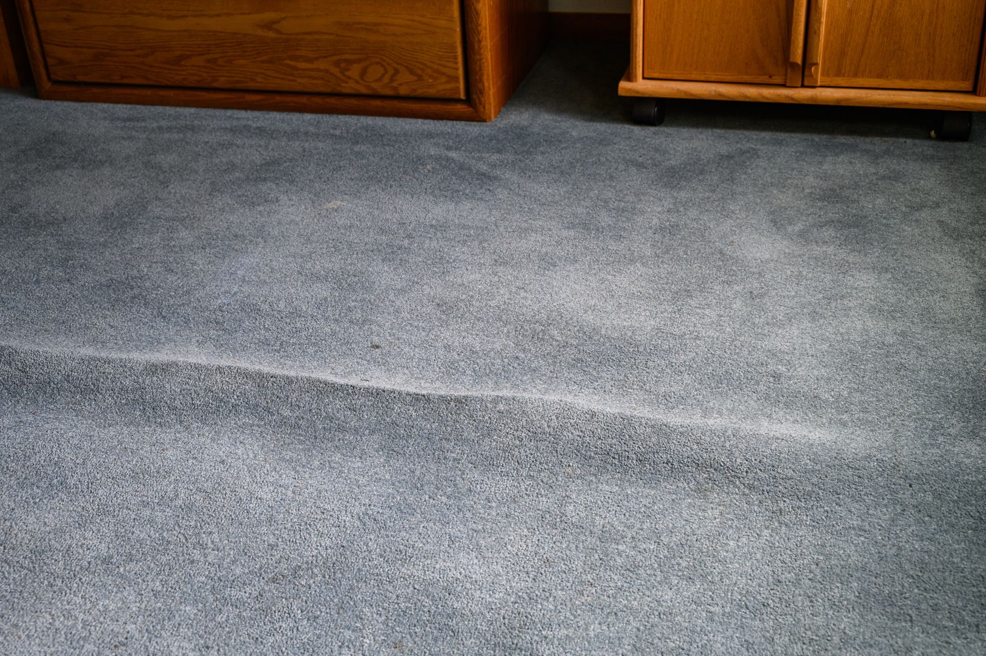 Blue carpet with a visible seam and ripples. Two wooden dressers sit on the carpet in the background.