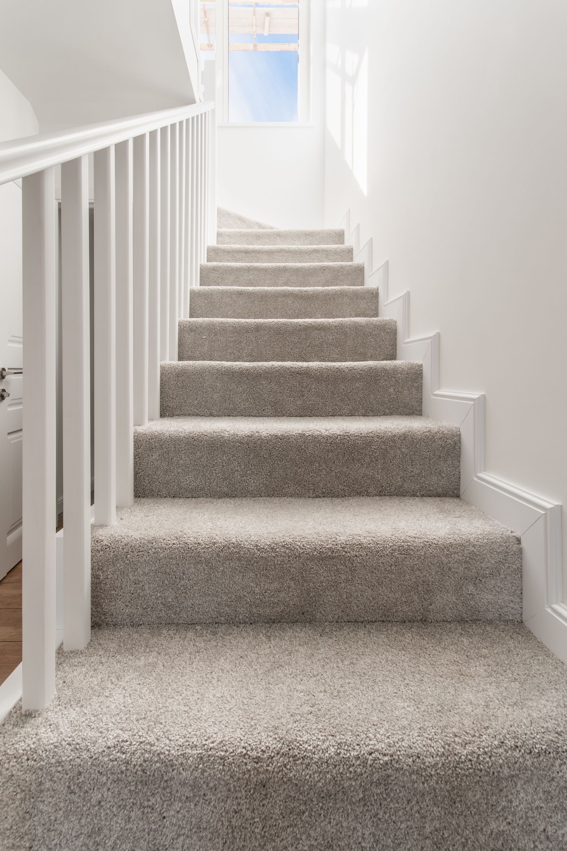 Staircase with light gray carpet and white railing, leading up to a window with a blue sky view.