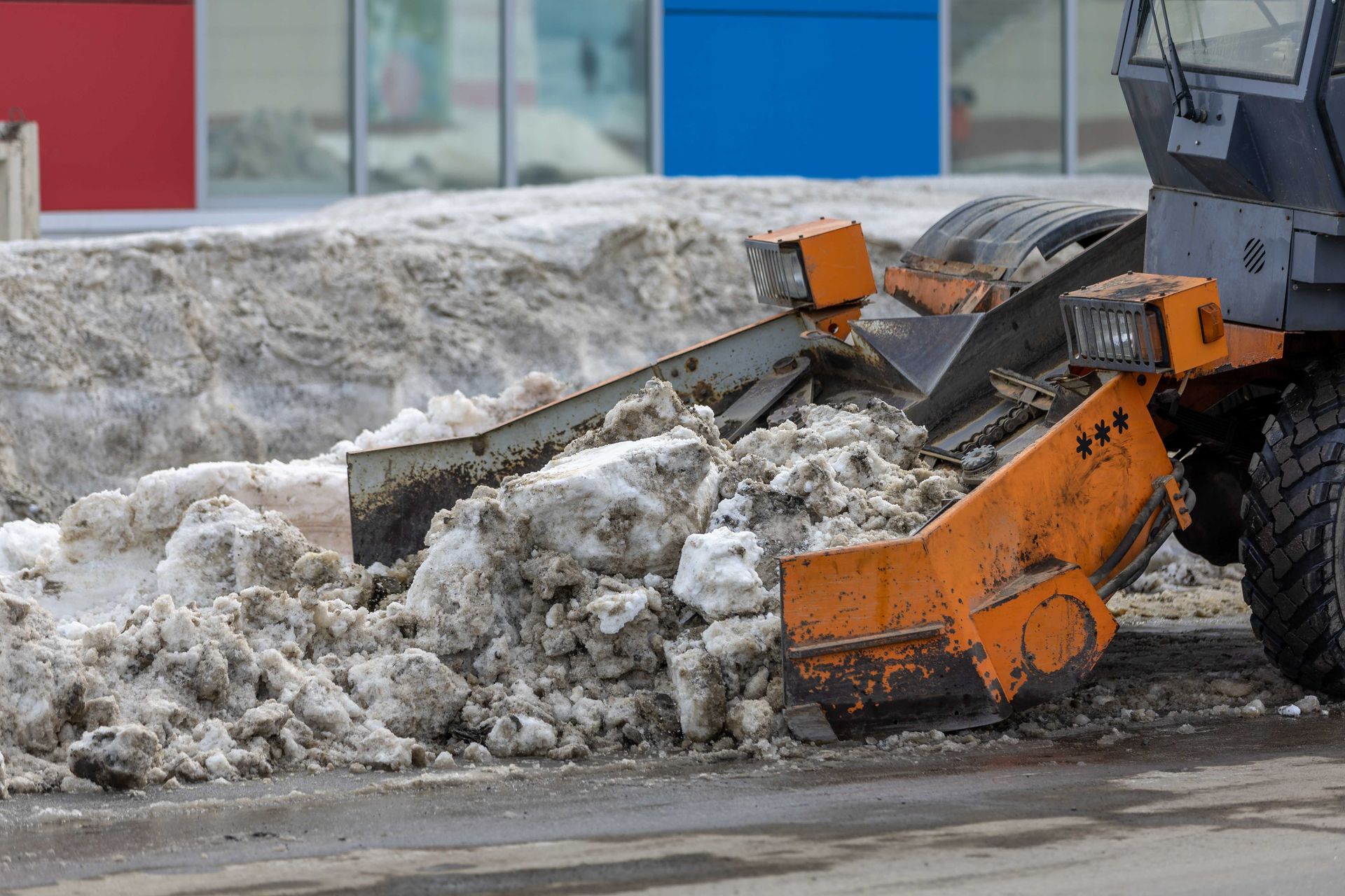 Orange snowplow clearing dirty snow from a roadside near colorful buildings