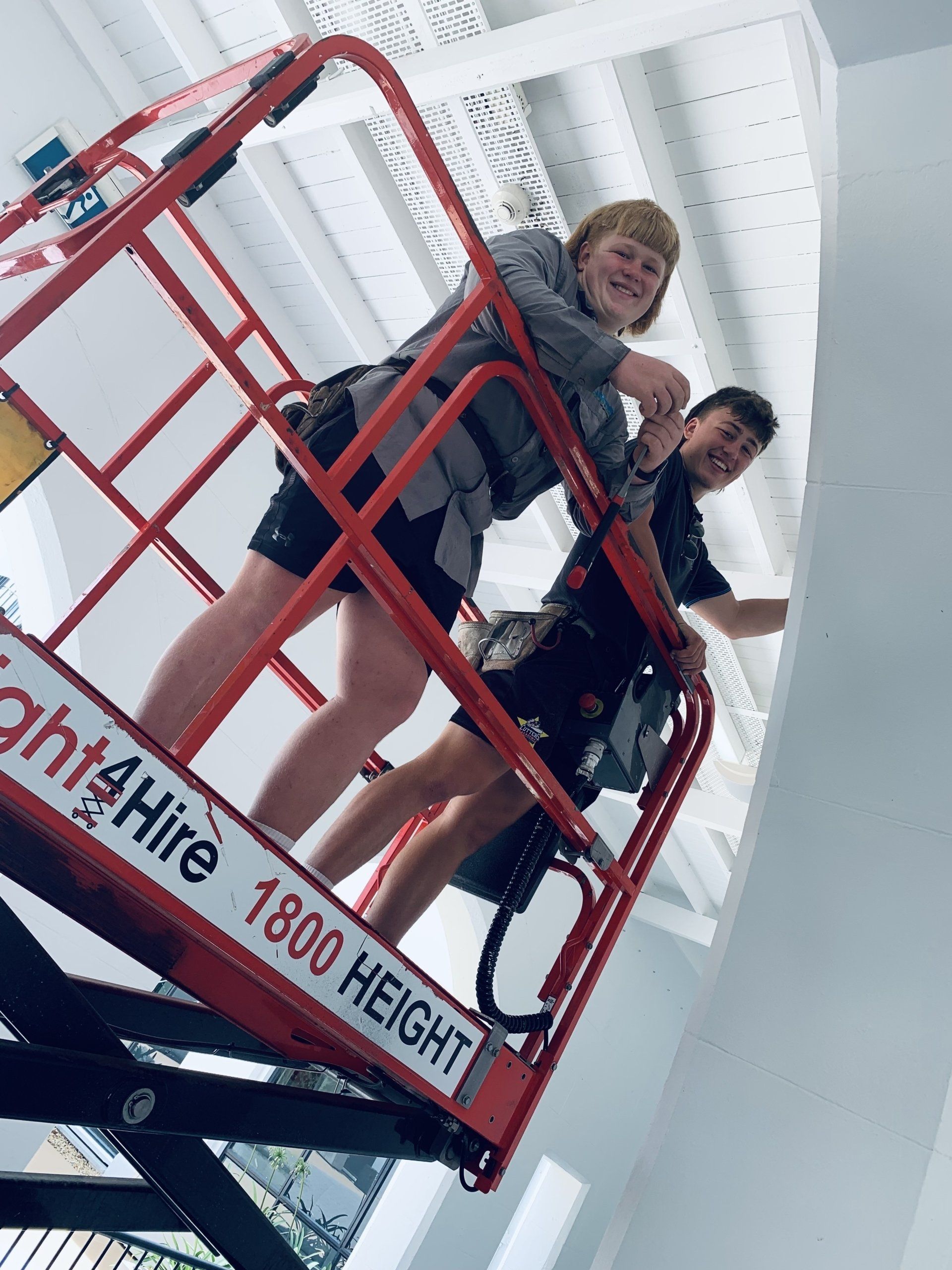 Two people on a red lift painting a white ceiling. One smiles, the other reaches towards the ceiling.