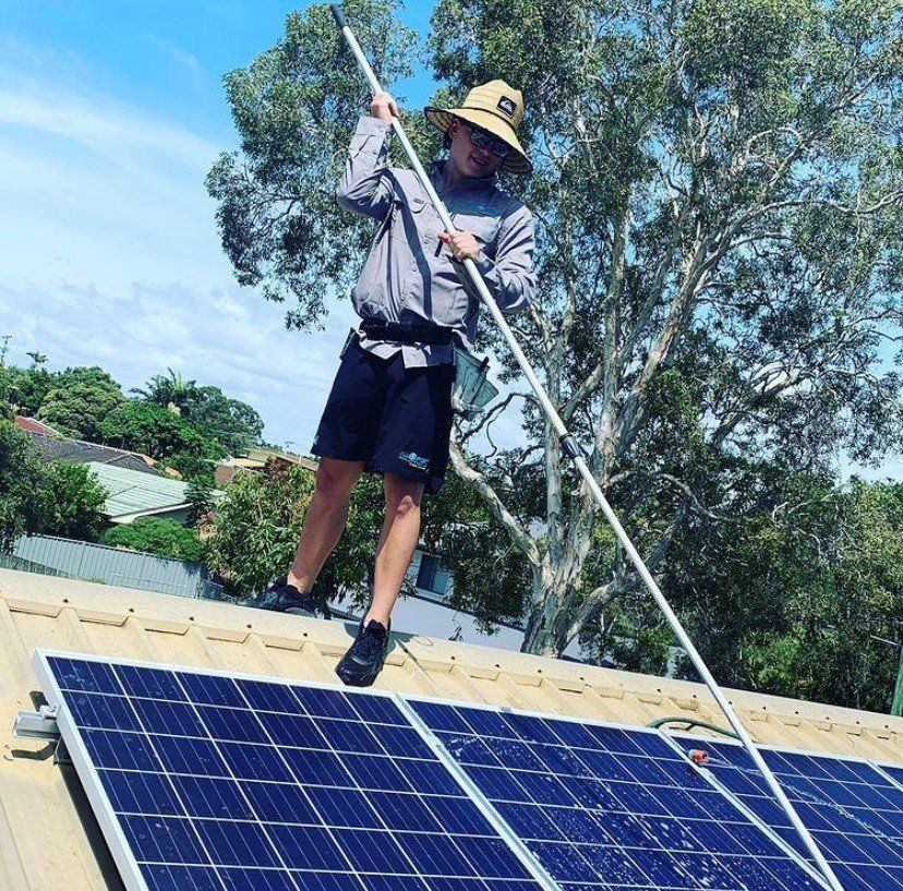 Person cleaning solar panels on a roof with a long pole under a sunny sky.
