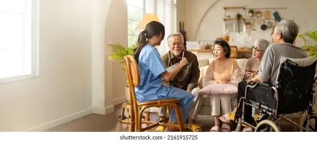 A caregiver talks with a group of seated seniors in a sunny room, one in a wheelchair.