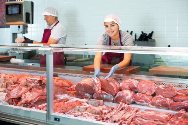 Butchers in a meat shop, one smiles while handling cuts of meat behind a glass display.