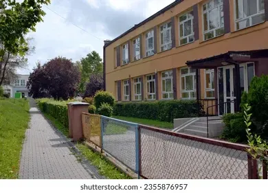 School building with tan exterior, sidewalk, and fenced-in area. Green bushes line the building and sidewalk. Overcast sky.