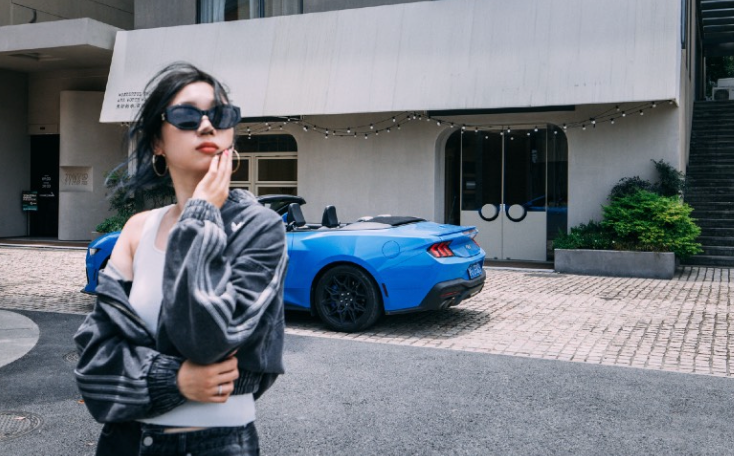 Woman in sunglasses poses by a blue convertible car in front of a white building.