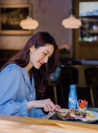 Woman at a table eating, a bottle of Oat beverage nearby. Indoor setting with soft lighting.