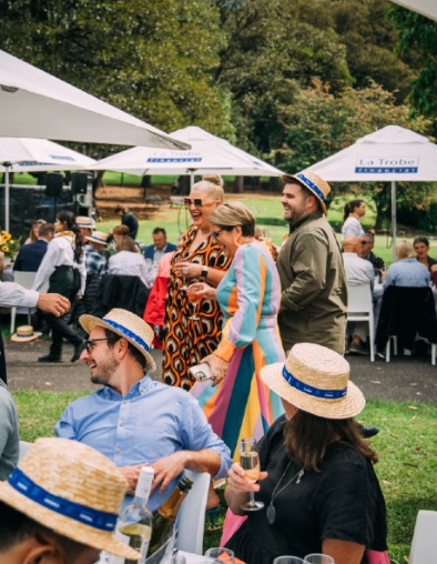 People in straw hats at an outdoor gathering, drinking, laughing under white umbrellas and trees.