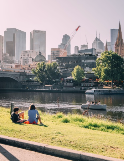 Two people on grassy bank near a river, with Melbourne city skyline in the background.