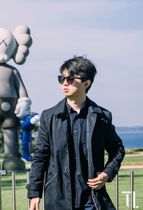 Man wearing sunglasses and a dark coat, standing outdoors near modern art sculpture.
