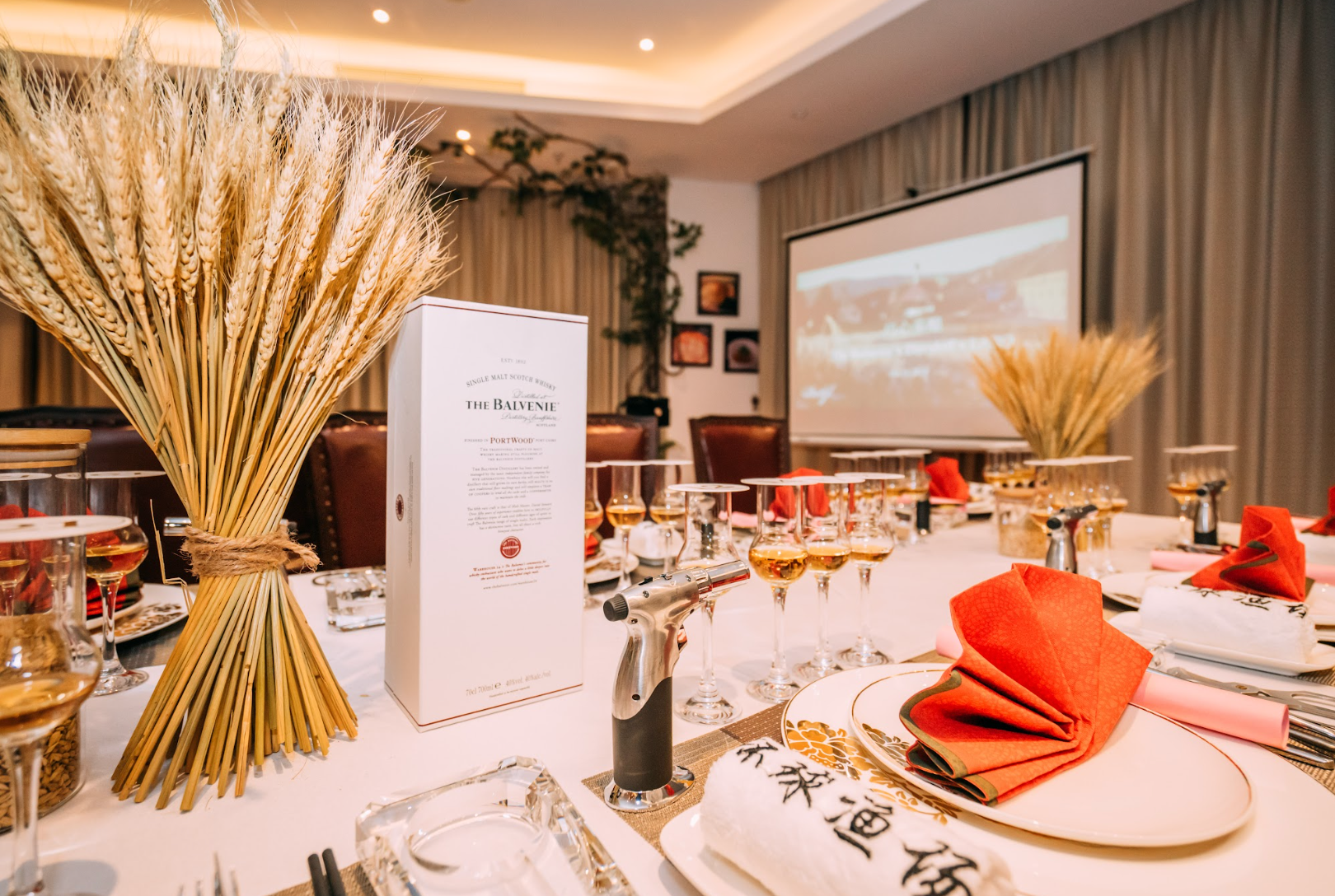 Table set for tasting event with whiskey box, wheat stalks, and a projector screen.