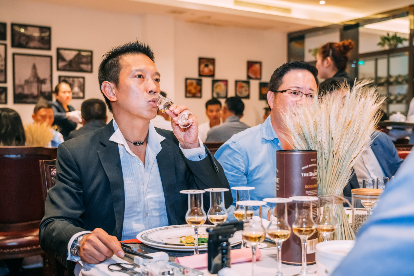 Man tasting drink at a table; other people and decorations in background.