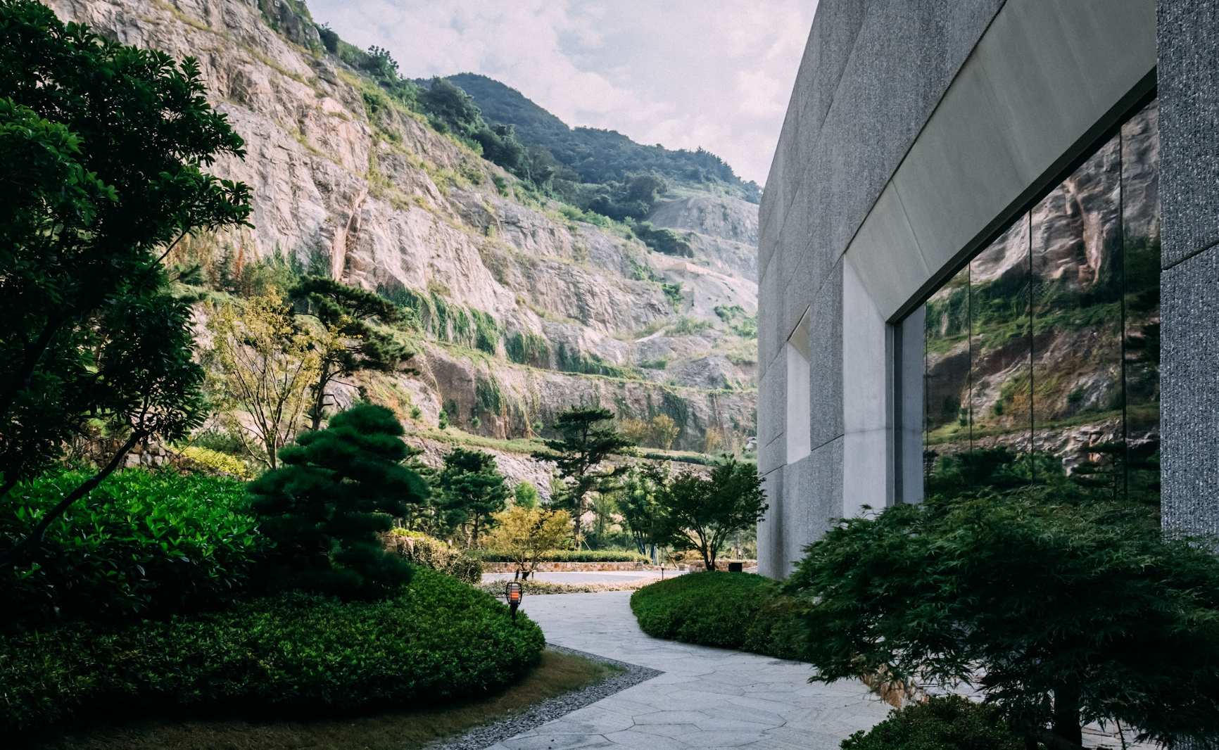 A modern building with a reflective glass wall beside a mountain with green foliage and a pathway.