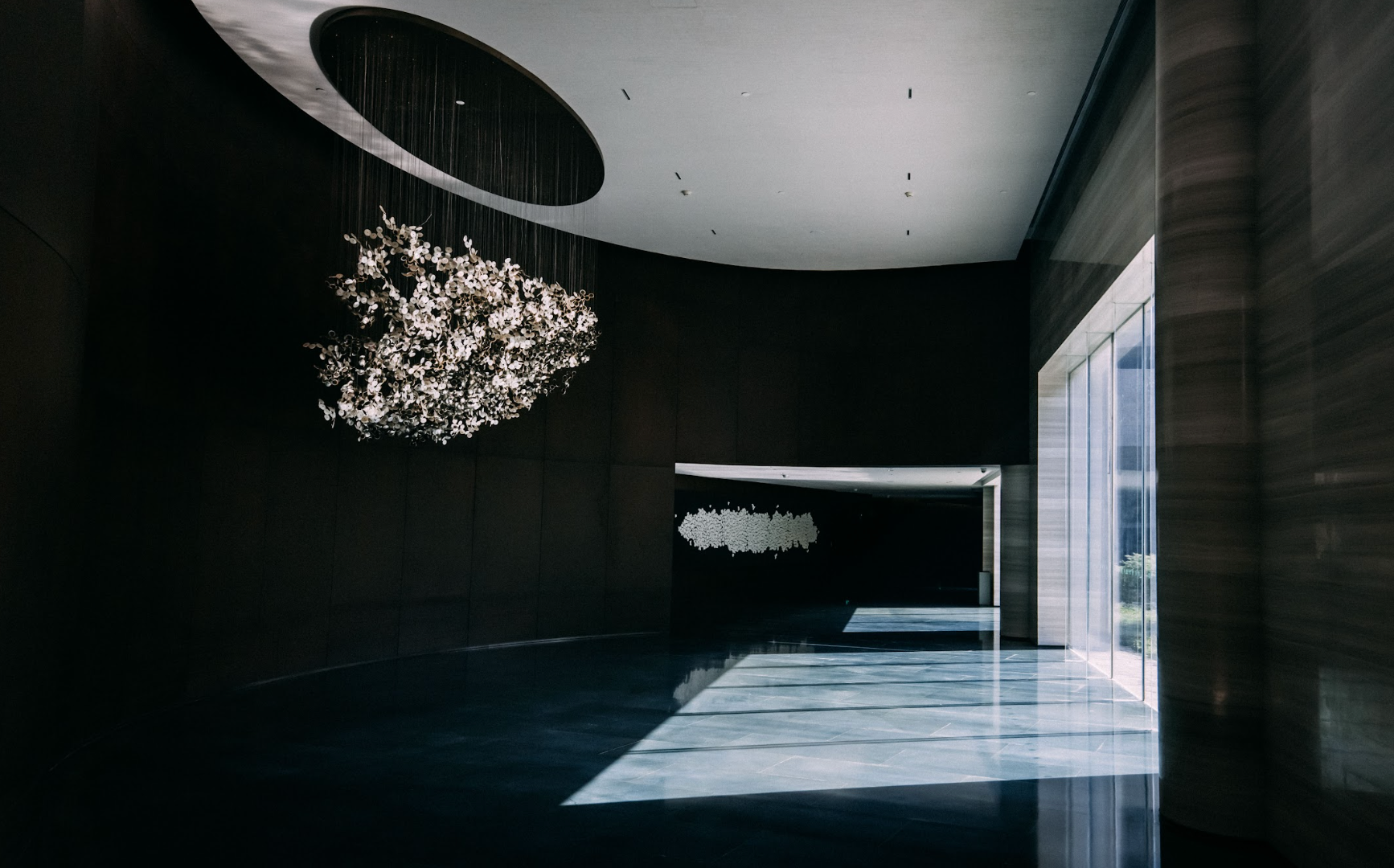 Dark interior with large light fixture, polished floors, and sunlight streaming through a glass door.