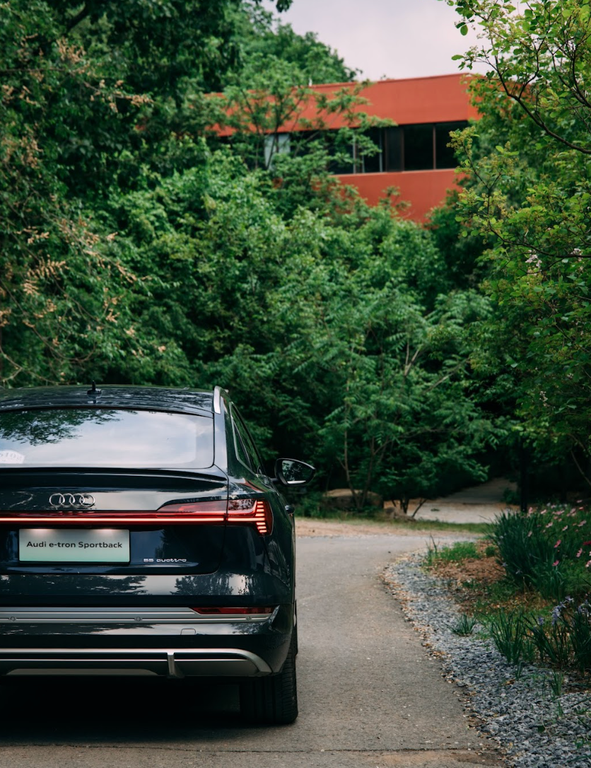 Black Audi SUV parked on a paved driveway with a red-roofed building and lush green trees in the background.