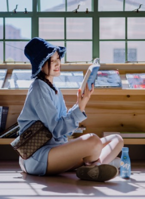 Woman reading a book, sitting on the floor in front of a window. Wearing blue hat, shirt, shorts, and a bag.