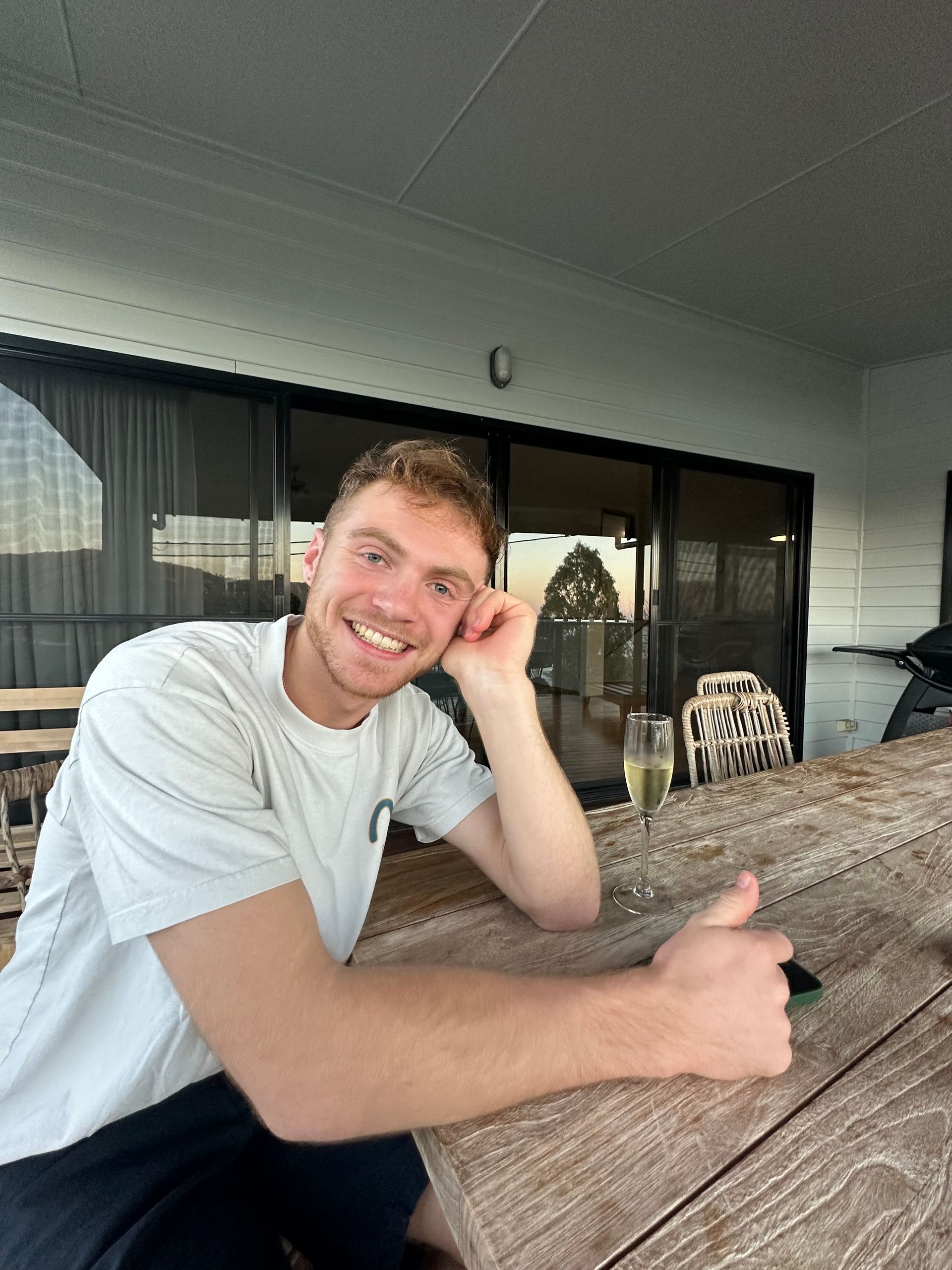 Man smiling, leaning on a wooden table outdoors, with a glass of champagne, and giving a thumbs-up.
