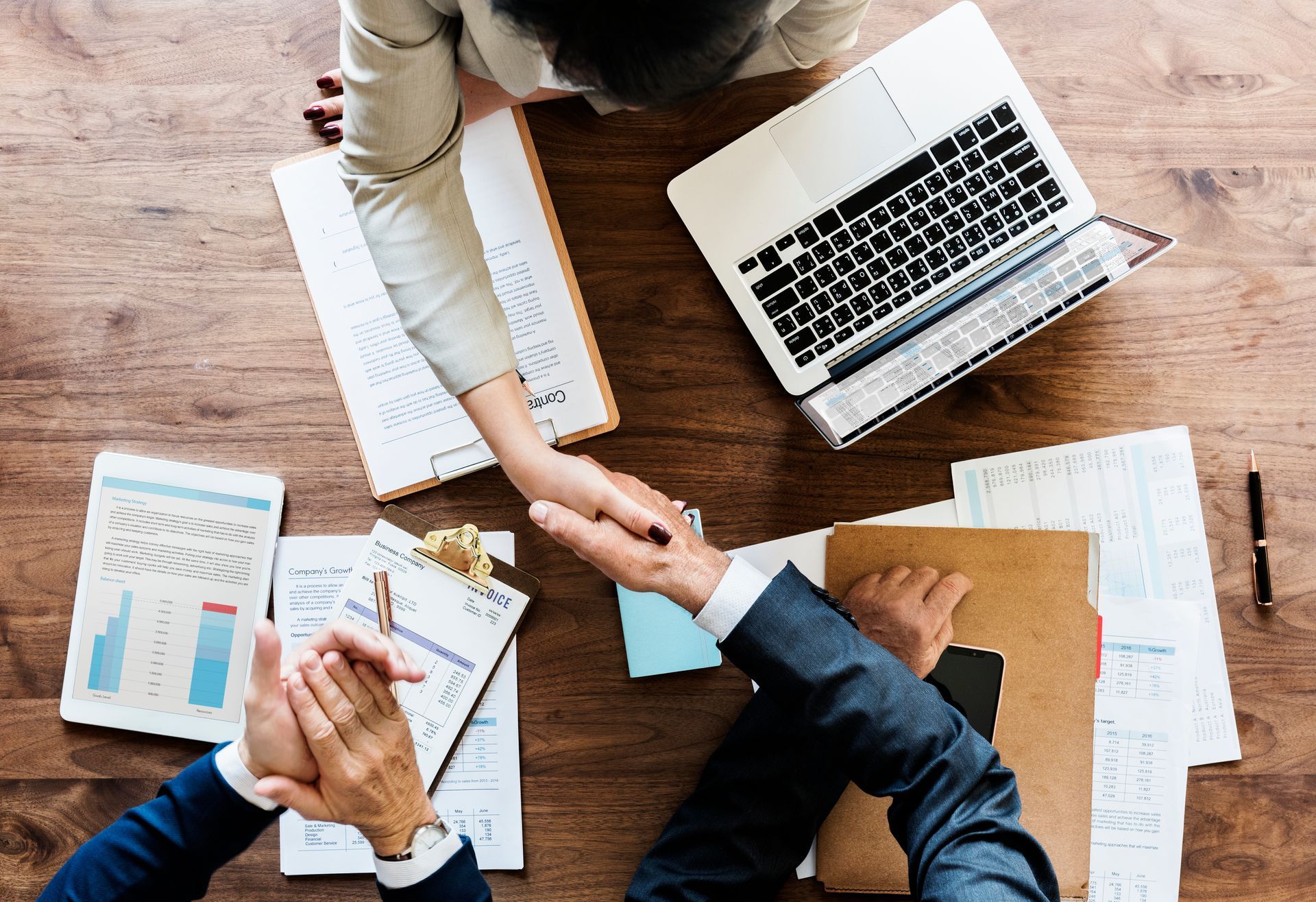 Three people in a meeting shake hands over a wooden table with documents and a laptop.