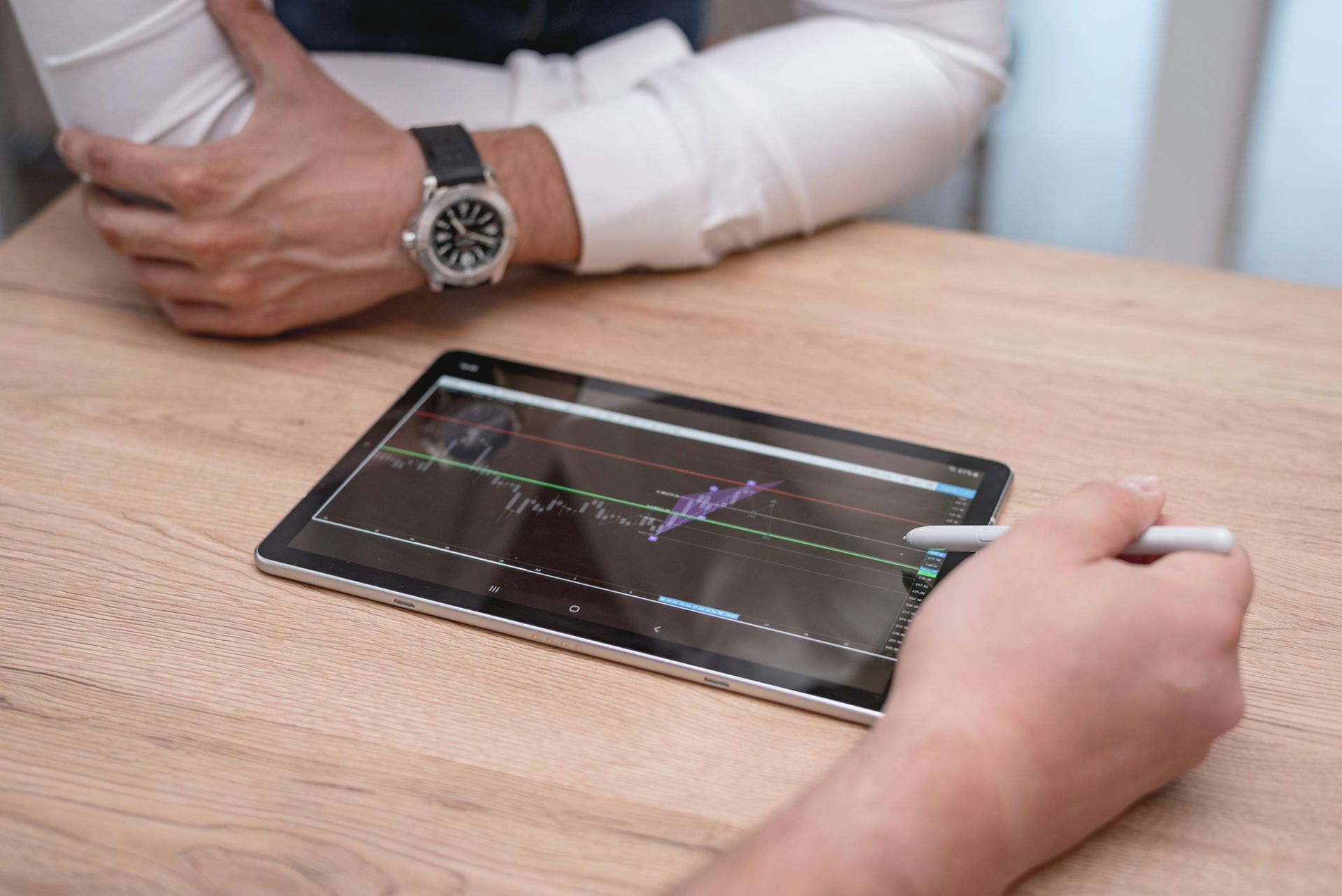 Hands using a stylus on a tablet displaying a financial chart on a wooden table