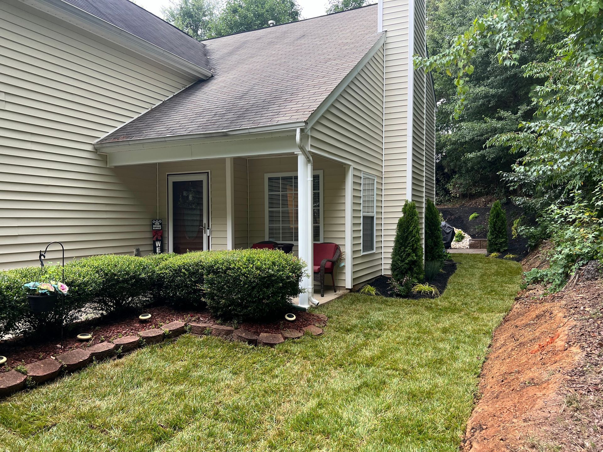 Front porch of a beige house with white columns, trimmed hedges, and a green lawn beside a wooded slope