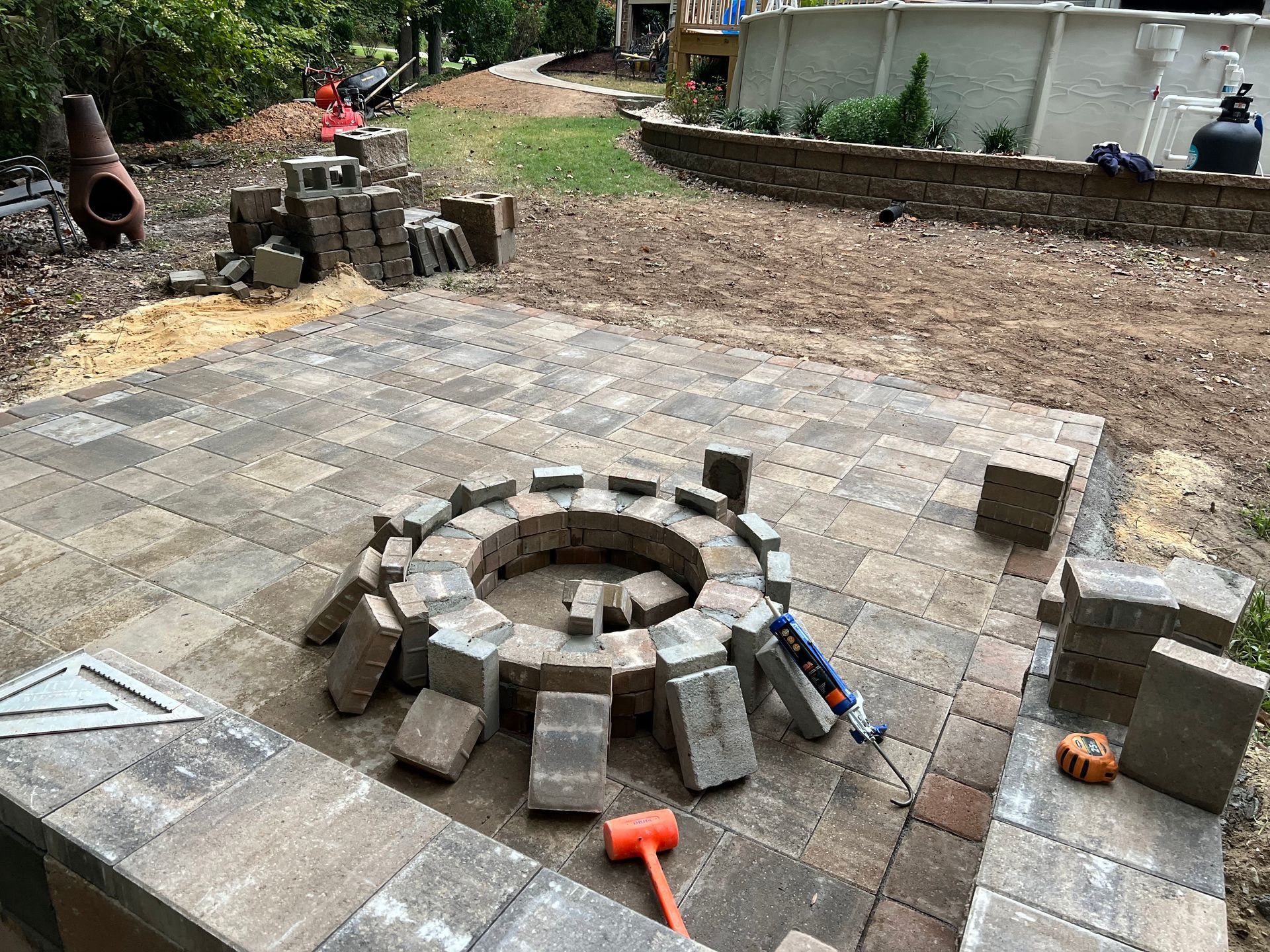 Backyard stone fire pit under construction with pavers, tools, and stacked blocks around it