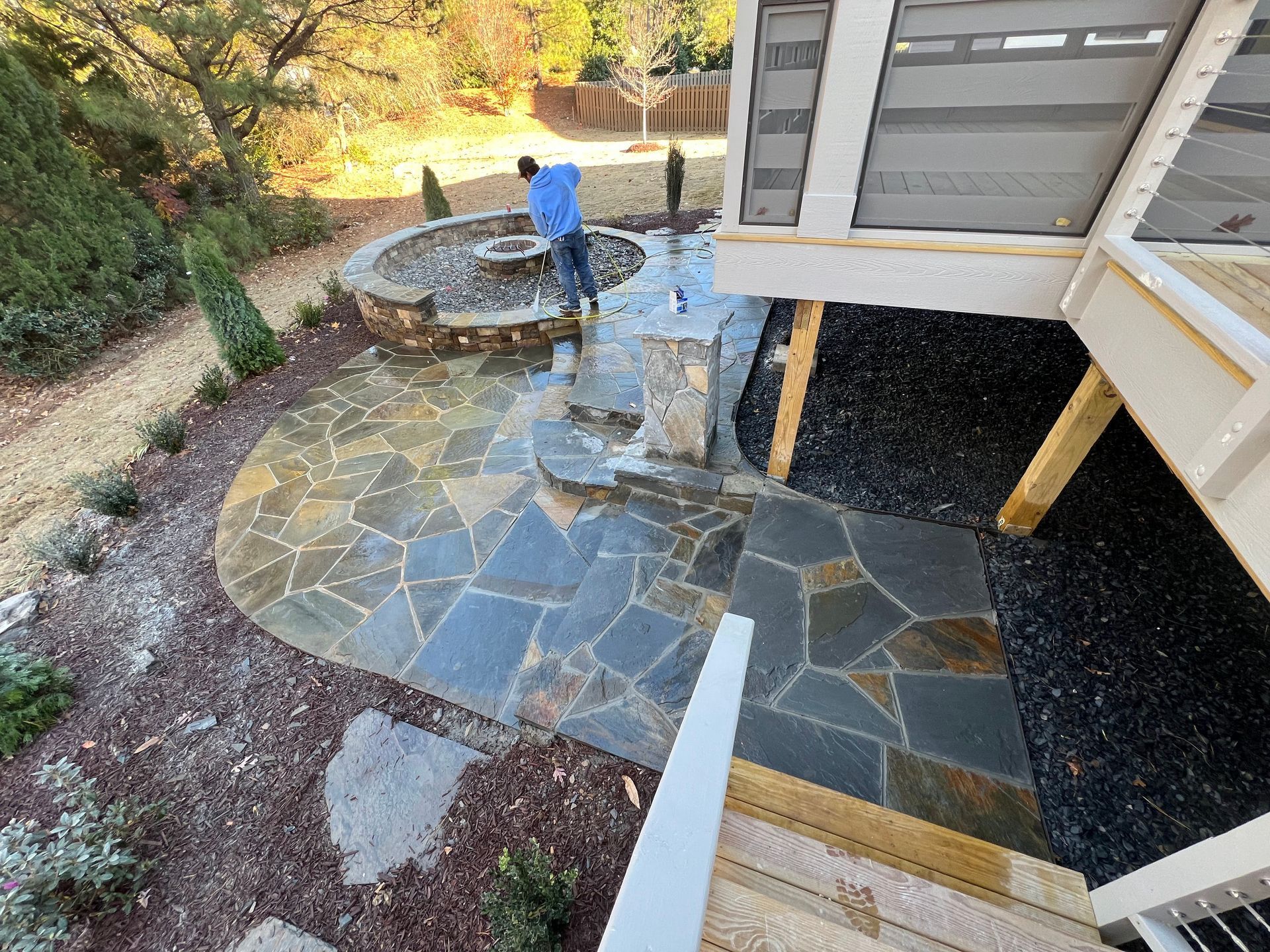 Stone patio and fire pit beside a house deck, with a person in a blue shirt standing near the wall.