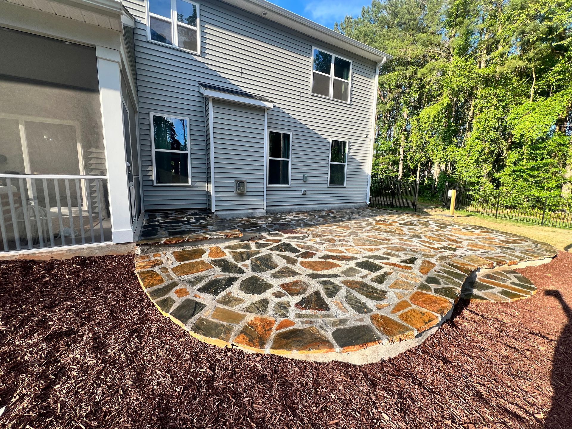 Stone patio with curved edge beside a house, bordered by dark mulch and trees in the yard