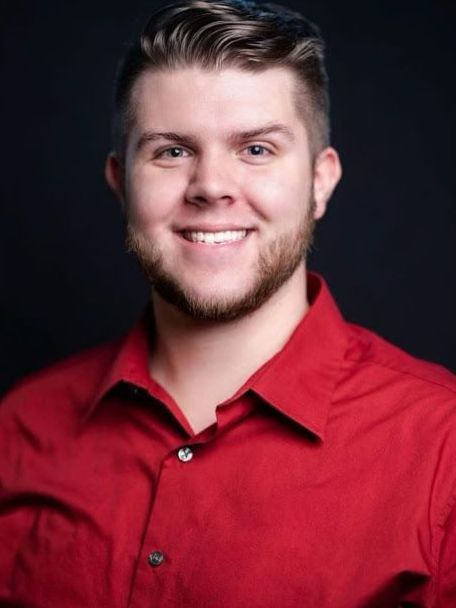 Man with a beard, smiling, wearing a red shirt, against a dark blue background.