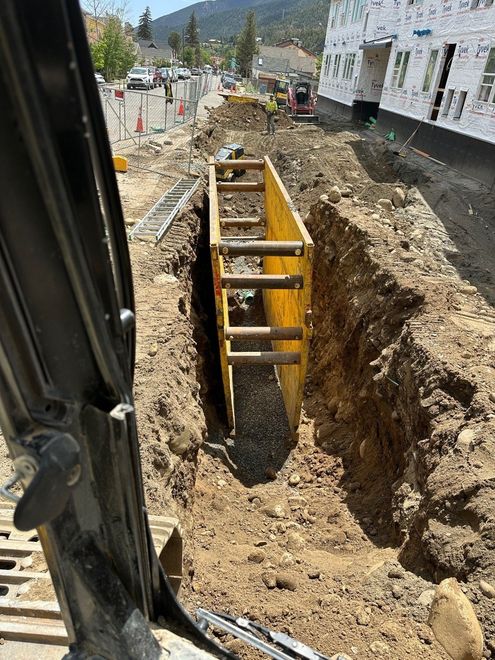 A yellow metal trench box sits inside a deep dirt excavation site next to a building under construction.
