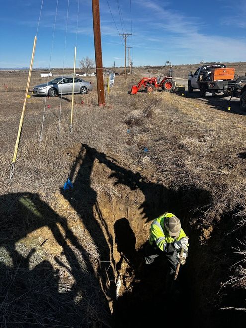 A worker in a high-visibility jacket repairs a utility line in an open trench near a parked truck and tractor.