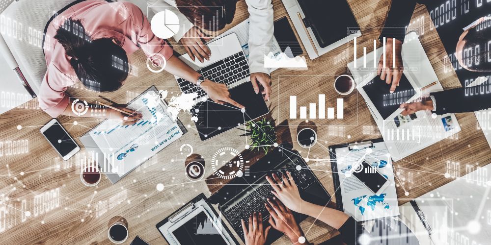 Business meeting with multiple people around a table, working on laptops and papers, with charts and graphs.