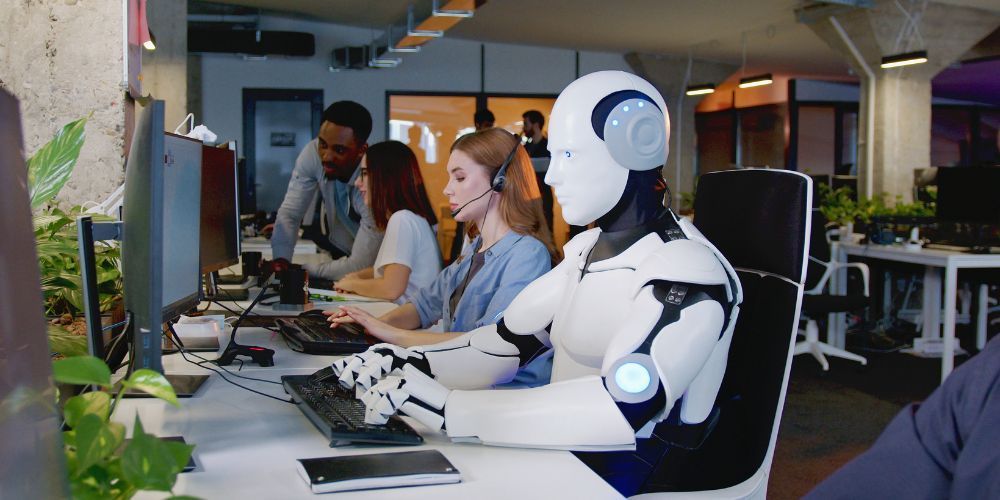 A robot is sitting at a desk in front of a computer.