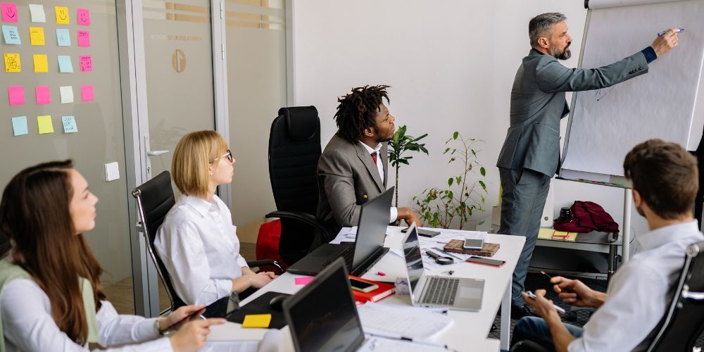 A man in a suit points at a whiteboard during a meeting with colleagues. Laptops and notes are on the table.