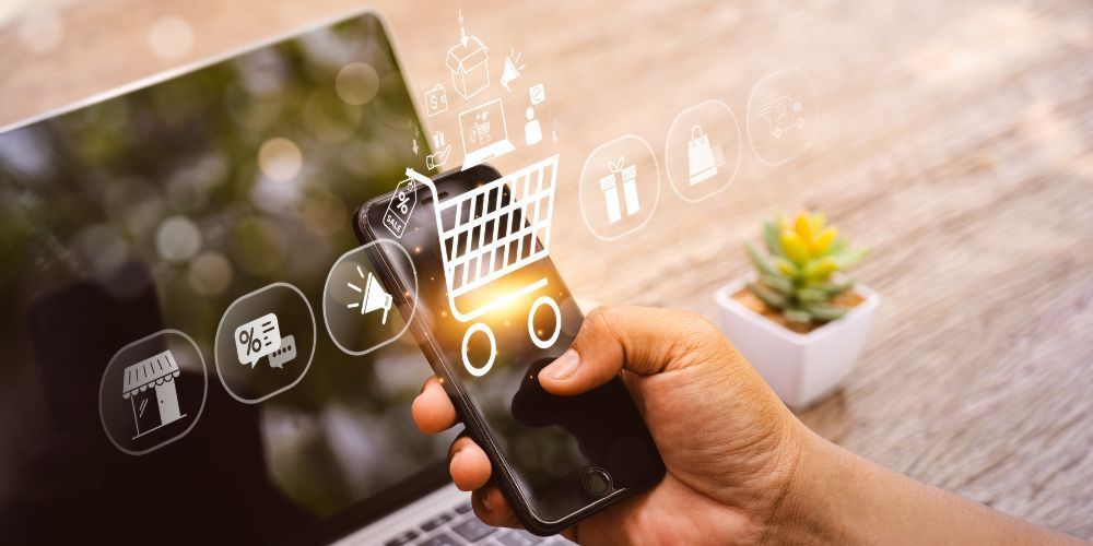 Person holding a smartphone with online shopping icons superimposed, near a laptop and small potted plant.