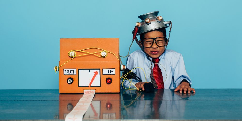 A person wearing a makeshift foil helmet and a red tie sits at a table next to an orange lie detector box with a gauge.