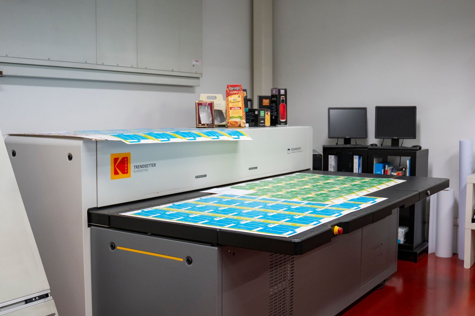Stampante di grande formato in una tipografia, che stampa fogli a colori accanto ai monitor dei computer.