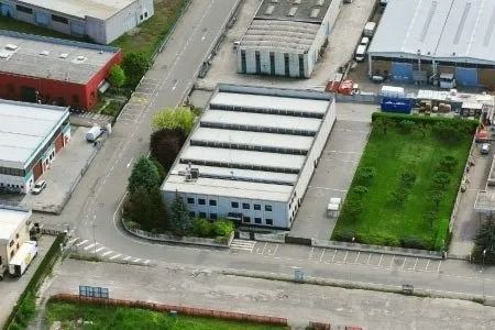 Veduta aerea di un edificio industriale con tetto bianco, circondato da strade, alberi e aree verdi.