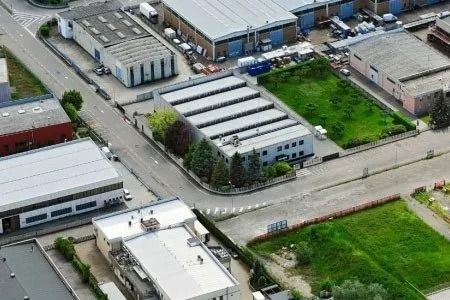 Veduta aerea di un'area industriale con magazzini, strade, edifici, camion parcheggiati e campi verdi.