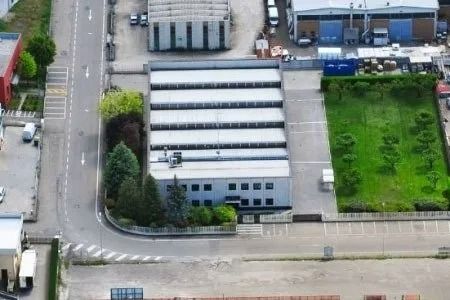 Veduta aerea di un magazzino industriale con tetti bianchi, situato accanto a una strada e a un prato verde.